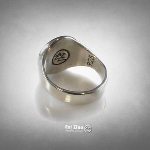 Op de afbeelding: Een zilveren ring met een getextureerd oppervlak en een 925 stempel aan de binnenkant. De ring heeft een gegraveerd logo aan de bovenkant.