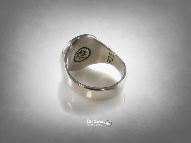 Op de afbeelding: Een zilveren ring met een getextureerd oppervlak en een 925 stempel aan de binnenkant. De ring heeft een gegraveerd logo aan de bovenkant.