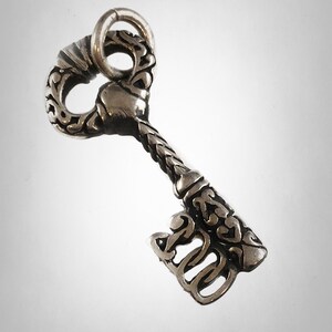 May include: A silver key charm with intricate details and a decorative design. The key has a heart-shaped bow and a detailed, ornate shaft.