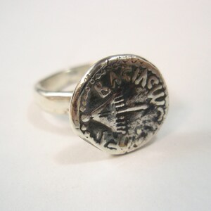 May include: A silver ring with a round, coin-shaped design featuring a menorah and Hebrew text. The ring is worn on a finger.