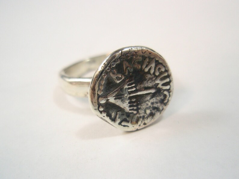 May include: A silver ring with a round, coin-shaped design featuring a menorah and Hebrew text. The ring is worn on a finger.
