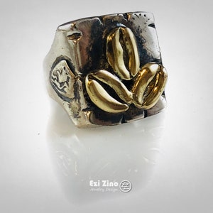 Puede incluir: Un anillo de plata con una cara cuadrada que presenta tres granos de café dorados. El anillo es texturizado y tiene un aspecto desgastado.