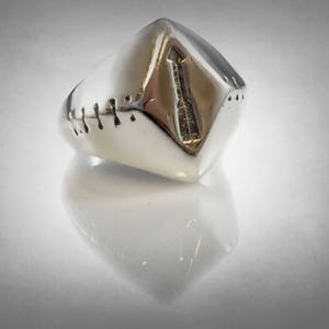 Puede incluir: Un anillo plateado con un dise&ntilde;o geom&eacute;trico. El anillo tiene una parte superior rectangular con un dise&ntilde;o grabado y peque&ntilde;as hendiduras en el lateral. El anillo es muy reflectante, con una superficie lisa y pulida.