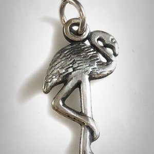May include: A silver charm in the shape of a flamingo. The flamingo is standing on one leg with its head bent down. The charm has a loop at the top for attaching to a bracelet or necklace.