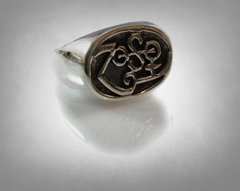 May include: A silver ring with an oval-shaped black and gold design. The design features a stylized letter "Z" with a swirl pattern.