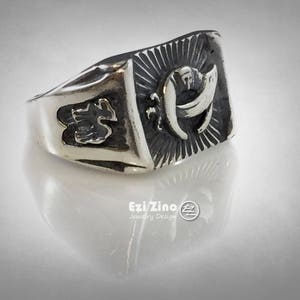 Puede incluir: Un anillo de plata con un dise&ntilde;o detallado que presenta una letra "H" estilizada y otros s&iacute;mbolos. El anillo tiene una superficie texturizada y una banda ancha.