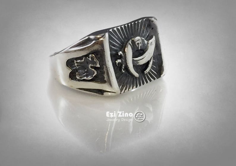 Puede incluir: Un anillo de plata con un dise&ntilde;o detallado que presenta una letra "H" estilizada y otros s&iacute;mbolos. El anillo tiene una superficie texturizada y una banda ancha.