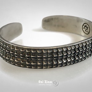 May include: A silver bracelet with a textured design. The bracelet is made of metal and has a square pattern. The bracelet is a simple design and would be a great addition to any jewelry collection.