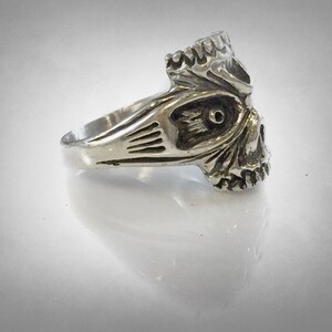 May include: A silver skull ring with intricate details. The skull has a detailed face with a prominent nose and eye sockets. The ring band is plain and smooth.