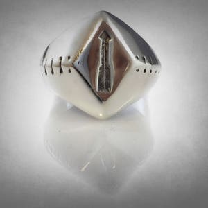 Puede incluir: Un anillo plateado con un dise&ntilde;o geom&eacute;trico. El anillo tiene forma de diamante con un dise&ntilde;o de flecha en el centro. Los lados del anillo tienen peque&ntilde;as hendiduras espaciadas uniformemente. El anillo es muy reflectante.