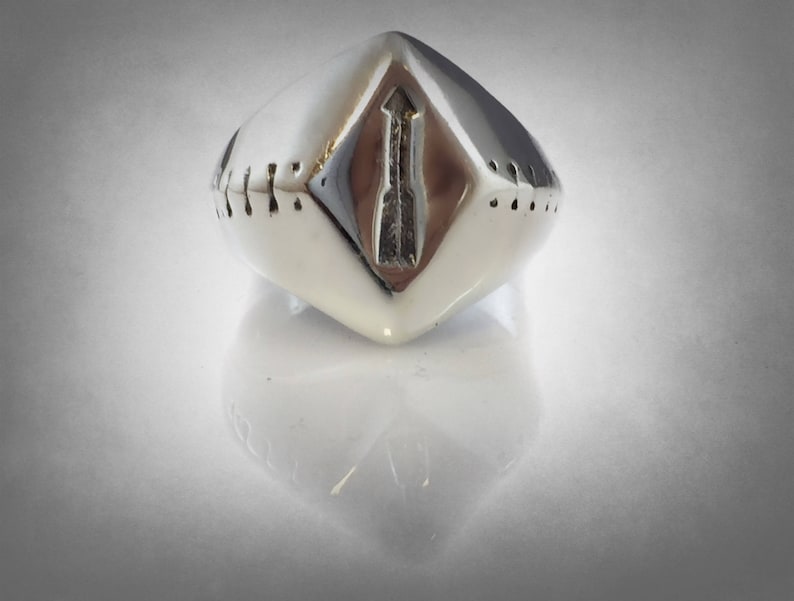 Puede incluir: Un anillo plateado con un dise&ntilde;o geom&eacute;trico. El anillo tiene forma de diamante con un dise&ntilde;o de flecha en el centro. Los lados del anillo tienen peque&ntilde;as hendiduras espaciadas uniformemente. El anillo es muy reflectante.