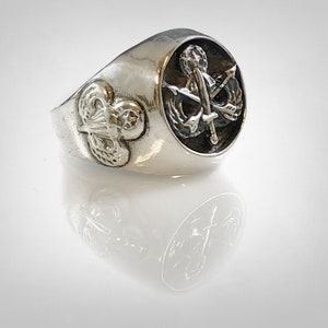 May include: A silver ring with a black enamel inlay featuring a military insignia. The insignia includes an anchor, sword, and wings.