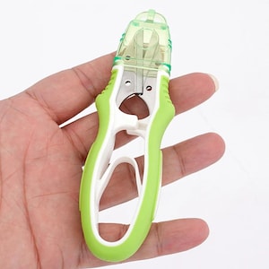 May include: A handheld thread cutter featuring a white and lime green plastic frame with a translucent green safety cap covering the sharp metal blades. The tool has an ergonomic design with a large finger loop and a spring-loaded mechanism.
