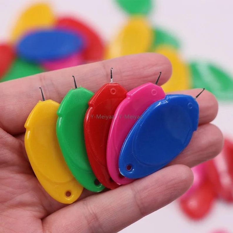 Puede incluir: Una mano sostiene una colecci&oacute;n de enhebradores de agujas de pl&aacute;stico de colores. Los enhebradores son amarillos, verdes, rojos, rosas y azules. Cada enhebrador tiene un peque&ntilde;o ojal y una aguja. M&aacute;s enhebradores en el fondo.