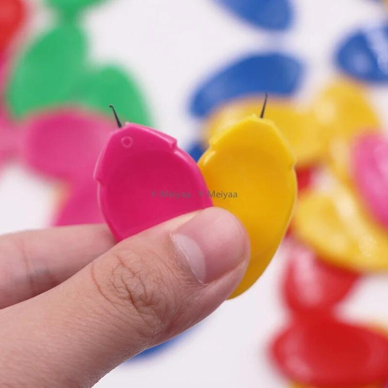 Puede incluir: Una mano sosteniendo dos enhebradores de costura de pl&aacute;stico de colores. Uno es rosa brillante, el otro es amarillo. Otros enhebradores en rojo, verde y azul est&aacute;n en el fondo. Cada enhebrador tiene una aguja peque&ntilde;a.