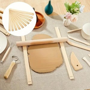 May include: A set of light-colored wooden clay rolling guides and tools, including a rolling pin, flat sticks with 5mm markings, a wire loop tool, a ribbon tool, and various wooden scrapers, arranged around a flattened piece of brown clay.