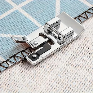 May include: A silver-colored metal sewing machine presser foot with a hinged design and a black plastic guide attachment. The presser foot features a polished finish and is positioned on a piece of fabric with a blue and white grid pattern.