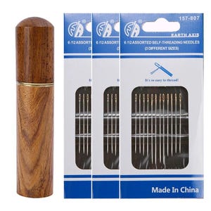 Op de afbeelding: Een houten naaldenkoker met een gouden band naast drie verpakkingen zelfrijgende naalden. De verpakkingen zijn wit en blauw met de tekst "EARTH AXIS" en "6/12 ASSORTED SELF-THREADING NEEDLES (3 DIFFERENT SIZES)". De tekst "It's so easy to thread!" is ook zichtbaar.