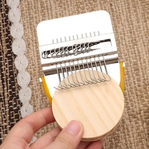 May include: A silver-colored weaving loom with a series of hooks and a wooden circular base. The loom has a rectangular top with evenly spaced vertical metal rods. The base is made of stacked, light-colored wooden circles.