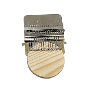 Könnte beinhalten: Ein Metallwebwerkzeug mit einer kreisförmigen Holzbasis. Das Werkzeug hat eine rechteckige Oberseite mit einer Reihe von Metallhaken und einer Reihe von Metallschlaufen. Ein gelbes Gummiband sichert die Basis. Das Werkzeug ist zum Weben bestimmt.