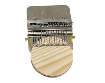May include: A metal weaving tool with a wooden circular base. The tool has a rectangular top with a series of metal hooks and a row of metal loops. A yellow rubber band secures the base. The tool is designed for weaving.