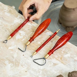 May include: A set of four pottery sculpting tools featuring polished reddish-brown wooden handles and brass ferrules. The tools include various metal wire loop ends in triangular, curved, and circular shapes for clay carving and detailing work.