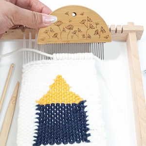 May include: A semi-circular bamboo weaving comb with a laser-engraved floral pattern and metal teeth, positioned over a small loom with white, yellow, and navy blue woven fabric, accompanied by two wooden weaving needles on the side.