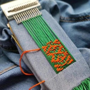 Może przedstawiać: Małe krosno z zieloną i pomarańczową przędzą tworzące tkany wzór na kawałku dżinsu. Krosno jest srebrno-białe, z wieloma metalowymi zębami. Dżins jest w kolorze jasnoniebieskim.