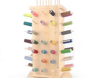 May include: A wooden thread spool organizer with multiple spools of colorful thread. The organizer is made of light-colored wood and has a rectangular shape with pegs to hold the spools. The spools are in various colors, including pink, green, blue, and yellow.