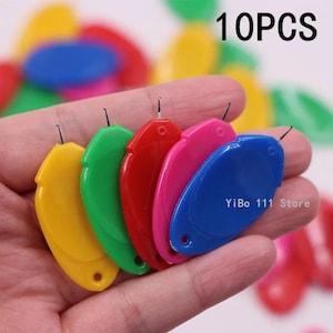 Puede incluir: Una mano sostiene cinco cortahilos de colores: amarillo, verde, rojo, rosa y azul. Cada cortahilos tiene una peque&ntilde;a cuchilla y una forma redondeada. El texto "10PCS" es visible arriba a la derecha, y "YiBo 111 Store" abajo.