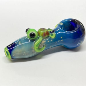 May include: A blue glass pipe with a green and brown turtle design. The pipe has a white speckled background and a blue bowl.