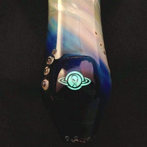 May include: A glass pipe with a blue, white, and green galaxy design. The pipe has a black mouthpiece and a green alien logo with a ring around it.