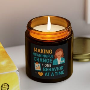 Può includere: Una candela accesa in un barattolo di vetro marrone con coperchio dorato. La candela ha un'etichetta nera con la scritta "MAKING MEANINGFUL CHANGE + ONE BEHAVIOR AT A TIME". Vicino c'è una piccola pianta grassa in vaso bianco.