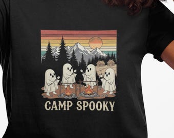 Może przedstawiać: Czarny t-shirt z retro grafiką przedstawiającą duchy biwakujące przy ognisku, z górami i drzewami w tle. Tekst "CAMP SPOOKY" znajduje się poniżej grafiki. Osoba na zdjęciu trzyma czarny smartfon.