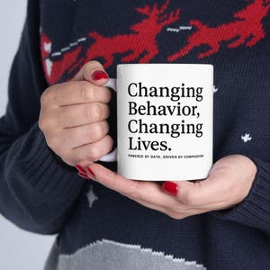 Op de afbeelding: Witte keramische mok met de tekst "Changing Behavior, Changing Lives." in het zwart. De mok heeft ook de tekst "POWERED BY DATA, DRIVEN BY COMPASSION" in een kleiner lettertype. De mok wordt vastgehouden door een persoon die een donkerblauwe trui draagt.