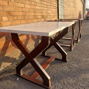 May include: A rectangular table with a light-coloured top and dark brown, crisscross-style legs. The table is made of wood and is positioned outdoors, leaning against a brick wall. The table's dimensions are approximately 183 cm long and 91 cm wide.