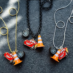 May include: Three necklaces with car-themed pendants. Each necklace features a different chain: gold, black, and silver. The pendants include a red car, an orange traffic cone, and a car wheel. The necklaces are displayed on a dark gray surface.