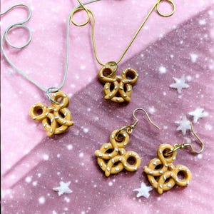May include: A collection of pretzel-shaped jewelry, including two necklaces and a pair of earrings. The pretzels are tan with white salt detailing. The necklaces have silver and gold chains. The background is pink with white star accents.