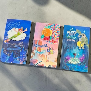 May include: Three rectangular display cards featuring enamel pins. The first shows Peter Pan with text "The Second Star to the Right". The second shows a house with balloons and text "Adventure is out there". The third shows a frog with text "You're never too old to wish upon a star".
