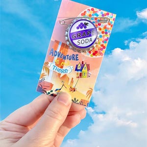 May include: A Grape Soda pin with a purple and white design, attached to a card with the text "Adventure is out there". The card features a colorful illustration of a house and balloons, set against a blue sky background.