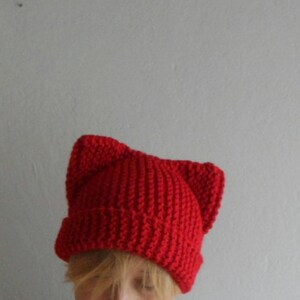 May include: A bright red knit hat with cat ears. The hat is made of a thick, textured yarn and features a ribbed brim. The cat ears are sewn on top, giving the hat a playful look. The hat is a solid color and appears to be handmade.