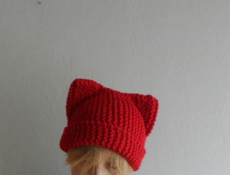 May include: A bright red knit hat with cat ears. The hat is made of a thick, textured yarn and features a ribbed brim. The cat ears are sewn on top, giving the hat a playful look. The hat is a solid color and appears to be handmade.