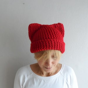 May include: A bright red knit hat with cat ears. The hat is made of a thick, textured yarn and is worn by a person wearing a white long-sleeved shirt. The hat's design features pointed ears at the top, adding a playful touch to the winter accessory.