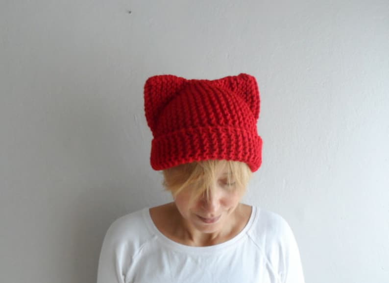 May include: A bright red knit hat with cat ears. The hat is made of a thick, textured yarn and is worn by a person wearing a white long-sleeved shirt. The hat's design features pointed ears at the top, adding a playful touch to the winter accessory.
