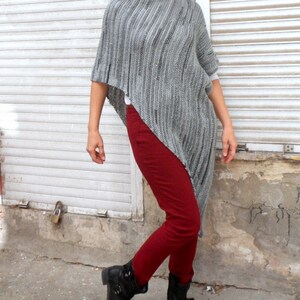 Pu&ograve; includere: Un poncho di maglia grigia con un orlo lungo e asimmetrico. Il poncho &egrave; indossato sopra pantaloni rossi e stivali neri.