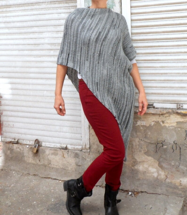 Pu&ograve; includere: Un poncho di maglia grigia con un orlo lungo e asimmetrico. Il poncho &egrave; indossato sopra pantaloni rossi e stivali neri.