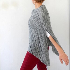 Pu&ograve; includere: Un poncho di maglia grigia con un orlo leggermente asimmetrico. Il poncho ha una trama a coste ed &egrave; indossato sopra una camicia bianca e pantaloni rossi.