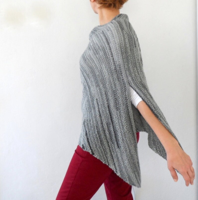 Pu&ograve; includere: Un poncho di maglia grigia con un orlo leggermente asimmetrico. Il poncho ha una trama a coste ed &egrave; indossato sopra una camicia bianca e pantaloni rossi.