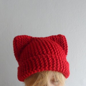 May include: A bright red knit hat with cat ears. The hat is made of a thick, textured yarn and has a ribbed brim. The cat ears are pointed and stand up from the top of the hat. The hat is a fun and playful accessory.