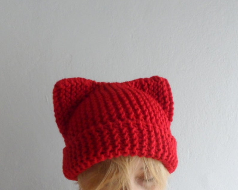 May include: A bright red knit hat with cat ears. The hat is made of a thick, textured yarn and has a ribbed brim. The cat ears are pointed and stand up from the top of the hat. The hat is a fun and playful accessory.
